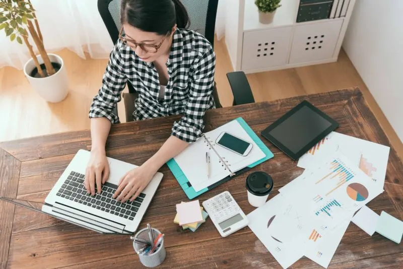 woman typing on laptop next to papers scattered on desk birds eye view